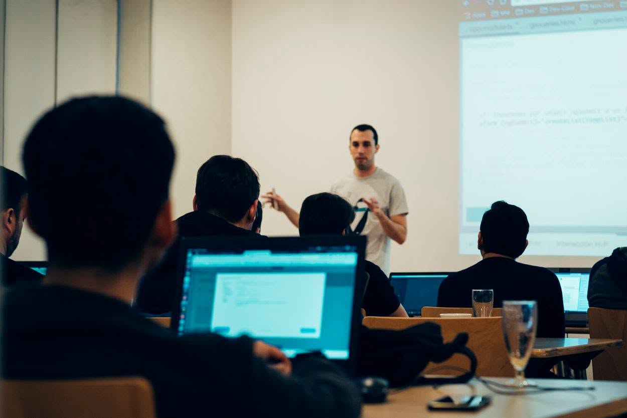Ein Mentor erklärt bei einem DevMeetings-Workshop vor der Gruppe, die Teilnehmenden arbeiten an Laptops